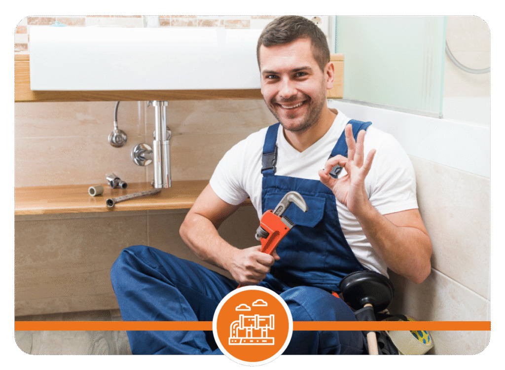 Plumber in blue overalls holding a pipe wrench and making an OK hand gesture while sitting on a wooden floor next to an open sink with plumbing tools.