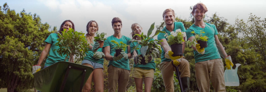 Group of five people in teal shirts holding plants, a watering can, and a wheelbarrow filled with greenery, standing in a garden with trees in the background.