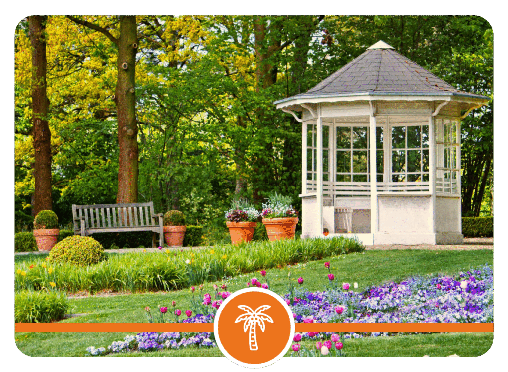 Garden with a wooden bench, two potted plants, white gazebo with windows and a shingled roof, vibrant flower beds, and lush trees in the background.
