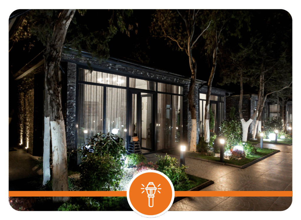 Modern building with large windows, white-trunked trees, flowering shrubs, and landscape lighting illuminating a paved walkway at night.