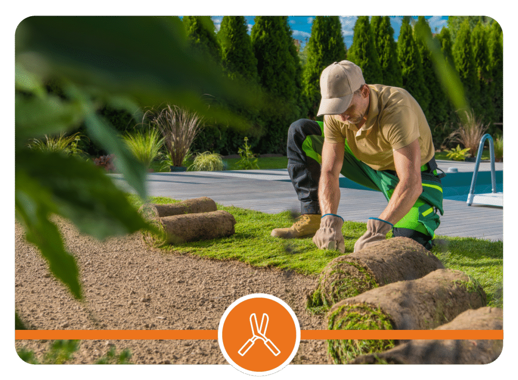 Person laying sod rolls on prepared soil beside a pool with surrounding plants, trees, and wooden deck in a landscaped backyard.