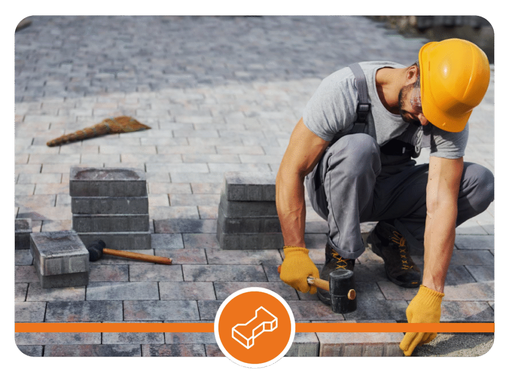Construction worker wearing yellow hard hat and gloves, kneeling on a paved surface while laying bricks with stacks of bricks, a trowel, and hammer nearby.